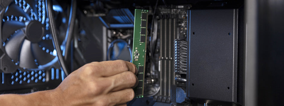A person's hand is holding a green RAM memory card in front of a computer's motherboard.