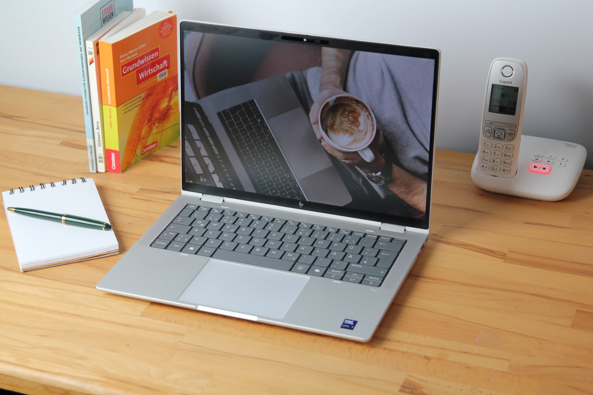 a laptop with a coffee cup on the screen, a notebook, a pen, and a phone on a wooden desk.