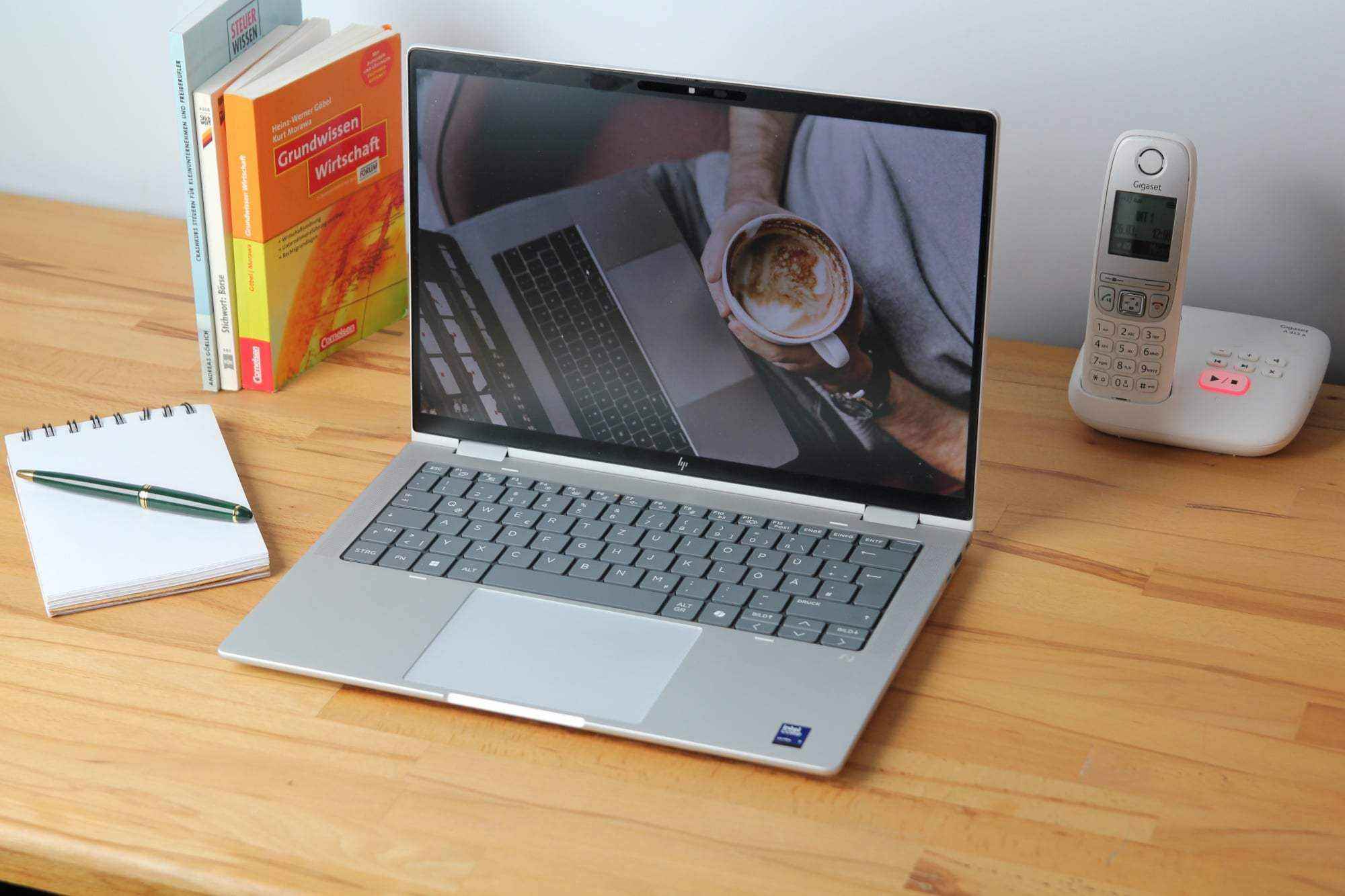 a laptop with a coffee cup on the screen, a notebook, a pen, and a phone on a wooden desk.